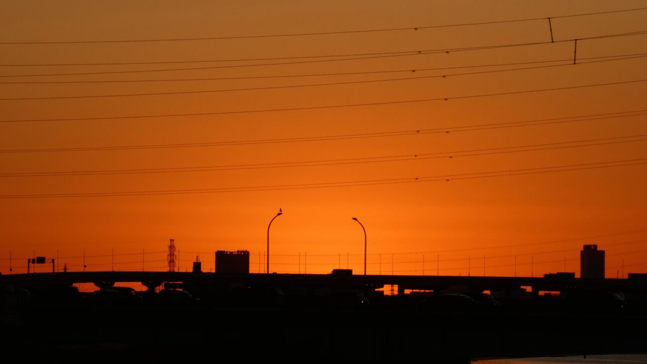 Silhouette of a modern city skyline, bridge, and streetlights against an intensely bright orange and red sunset or sunrise glow, symbolizing the contrast between the dark world and the shining light of faith.